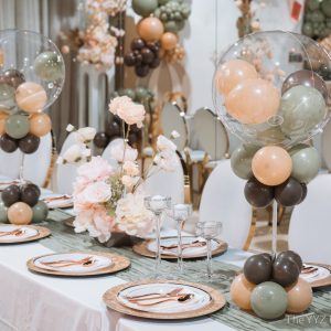 Guest table styled for a travel-themed baby shower at Styld Spaces Events Studio in Scarborough. Clear bubble balloon centerpieces in sage, peach and brown rise above gold charger plates, rose gold cutlery, blush rose florals and a soft sage table runner.