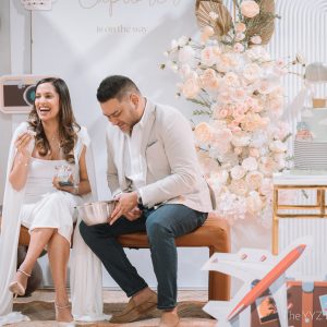 The parents-to-be laugh together during a baby shower game beside a two-tier travel-themed cake, a blush floral installation and an organic balloon garland at their 'Little Explorer' baby shower, Styld Spaces Events Studio, Scarborough.