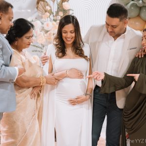 The expecting mother in a white gown surrounded by smiling family members celebrating at a travel-themed baby shower in front of a blush floral and balloon backdrop at Styld Spaces Events Studio, Scarborough, Toronto.