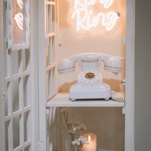 A white vintage-style telephone audio guestbook inside a phone booth prop, lit by a glowing 'Ring Ring' neon sign with a 'The Keepsake Line' card, letting guests record voice messages at a travel-themed baby shower, Styld Spaces Events Studio, Toronto.