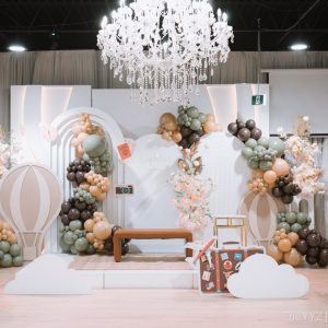 Wide view of a travel-themed 'A little Explorer is on the way' baby shower at Styld Spaces Events Studio in Scarborough, Toronto. The setup features a sage green, tan and brown organic balloon garland, hot air balloon cutout props, cloud and suitcase decor, blush floral arrangements, a crystal chandelier and gold-accented guest chairs.